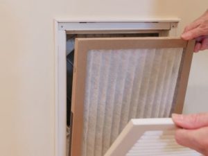 Hands removing a white air filter from a wall vent to improve indoor air quality at home. Air Works Heating & Air services.