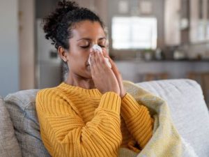 Woman in a cozy indoor setting, wearing a yellow sweater, sits on a couch with tissues and a blanket.