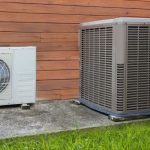 Two air conditioning units, one white and smaller, one larger and grey, positioned against a wooden wall, on concrete ground.