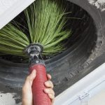 Hand holding a brush cleaning tool for HVAC duct maintenance with a tire in the background.