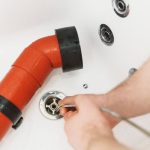Person's hand using a wrench on a red pipe in a bathtub for Air Works Heating & Air plumbing maintenance service.