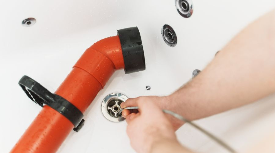 Person's arm using flexible hose and orange pipe for plumbing maintenance near circular drain in bathtub setting.