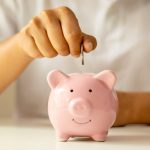 Hand inserting a coin into a pink piggy bank, symbolizing savings and cost reduction in HVAC expenses.